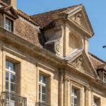 Bâtiment en pierre avec filets et pics anti-pigeons, oiseaux volant devant la façade sous un ciel bleu.
