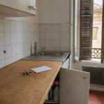 Cuisine d'un appartement ancien, placard sous l'évier entrouvert, trousseau de clés sur le plan de travail et un cafard sur le carrelage