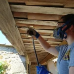 Intervenante en masque respiratoire pulvérisant un produit de traitement sous les chevrons d'un avant-toit en bois